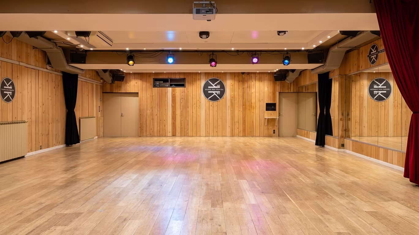 Grande salle de 150 m² du Studio des Rigoles, vide : parquet clair, murs lambrissés de pin aux médaillons du logo de l’école, rideaux noirs, rampe de projecteurs colorés, rideau rouge devant les miroirs.